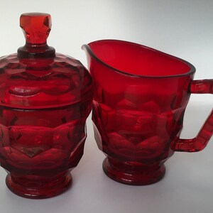 May include: A red glass creamer and sugar bowl set with a faceted design. The creamer has a handle and the sugar bowl has a lid.