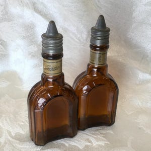 May include: Two vintage brown glass bottles with metal caps. The bottles have a unique shape and a label that reads "Stetson". The caps are a dull silver color. The bottles are displayed on a white, textured surface.