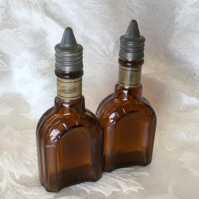 May include: Two vintage brown glass bottles with metal caps. The bottles have a unique shape and a label that reads "Stetson". The caps are a dull silver color. The bottles are displayed on a white, textured surface.