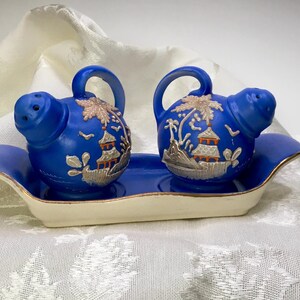 May include: A pair of cobalt blue salt and pepper shakers with a matching tray. Each shaker has a handle and a decorative silver and gold design of a pagoda and landscape. The tray is off-white with a gold trim.