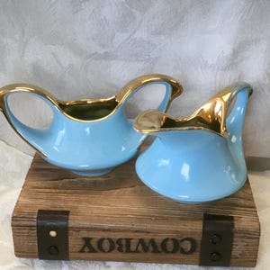 May include: Two light blue ceramic pitchers with gold-toned rims and handles. The pitchers feature a smooth, glossy finish and curved silhouettes. One pitcher has a double-handle design, while the other is a single-handled creamer style vessel.