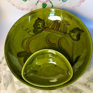May include: A set of two green ceramic serving bowls. The larger bowl has a wide rim and is decorated with a floral design in brown and black. The smaller bowl is a rounded triangular shape. The bowls are on a white surface.