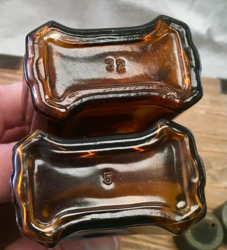 May include: Two amber-colored glass bottles, viewed from the bottom, with a rectangular base and rounded edges. One bottle has the number "32" embossed, the other has "5". The bottles are empty.