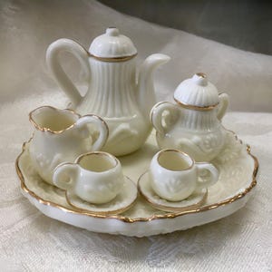 May include: A miniature cream-colored tea set with gold trim. The set includes a teapot, sugar bowl, creamer, two teacups, and saucers, all arranged on a decorative tray. The pieces have a delicate, ornate design.