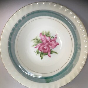 May include: A white ceramic bowl with a green and gold rim. The bowl features a pink orchid with green leaves in the center.