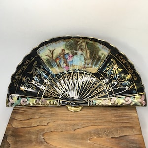 May include: A decorative fan with a black background and gold accents. The fan features a painted scene of people in a garden. The fan is resting on a wooden surface. The fan has a gold metal base.