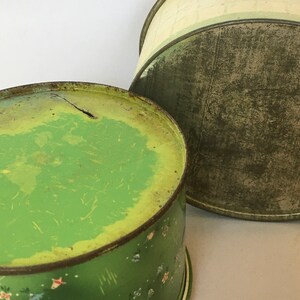 May include: Two vintage green metal tins with floral designs. The top of the tin on the left is green with a yellow center. The top of the tin on the right is green with a white center.
