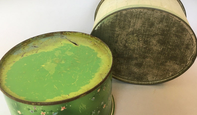 May include: Two vintage green metal tins with floral designs. The top of the tin on the left is green with a yellow center. The top of the tin on the right is green with a white center.