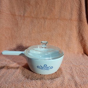 May include: A white saucepan with a clear glass lid and a light blue handle. The pan features a blue floral design on the side. The background is a peach-colored fabric.