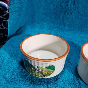May include: Two white ceramic ramekins with a brown rim and a colorful countryside scene painted on the side. The ramekins are on a blue towel.