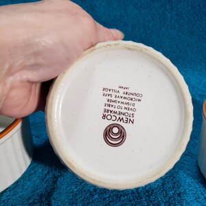 May include: White stoneware ramekins with brown trim. The ramekins are oven, dishwasher, and microwave safe. The text on the bottom reads "Newcor Stoneware Oven to Table Dishwasher Safe Microwave Safe Country Village Japan".