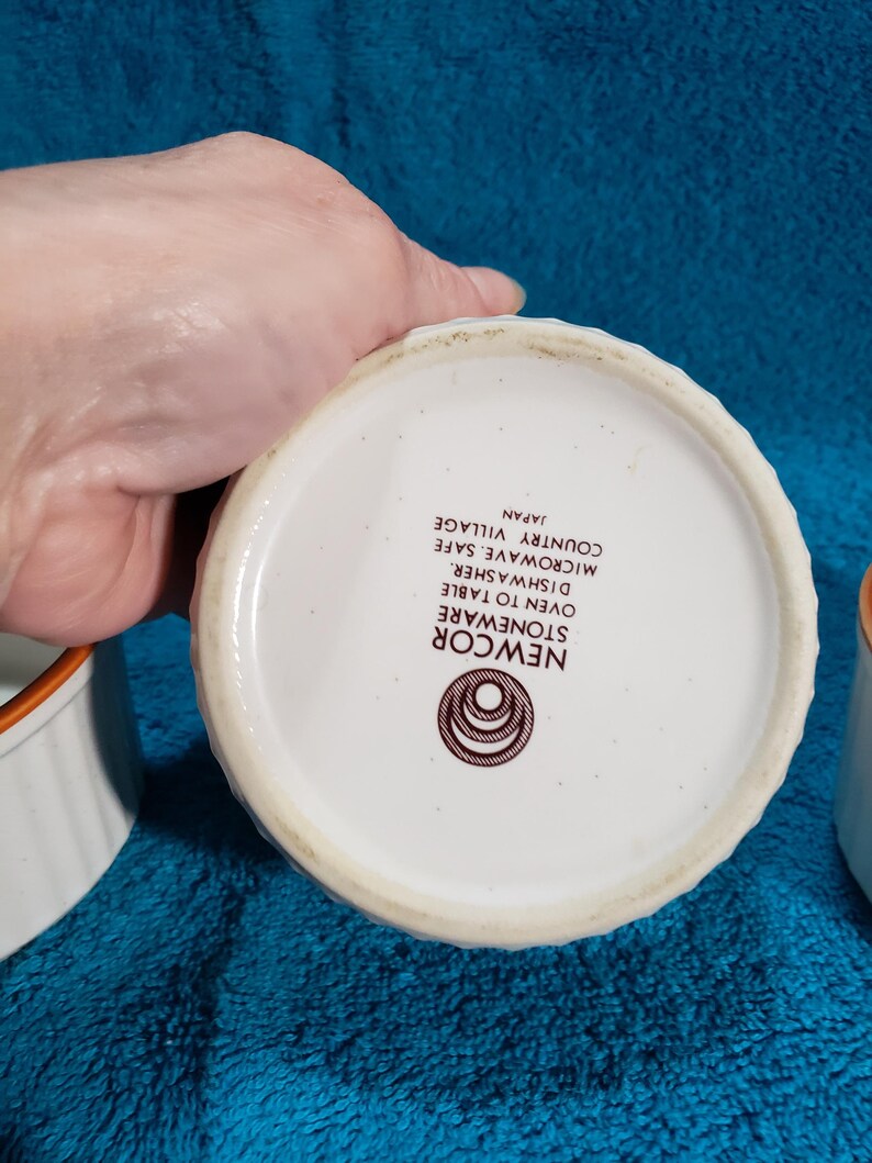 May include: White stoneware ramekins with brown trim. The ramekins are oven, dishwasher, and microwave safe. The text on the bottom reads "Newcor Stoneware Oven to Table Dishwasher Safe Microwave Safe Country Village Japan".