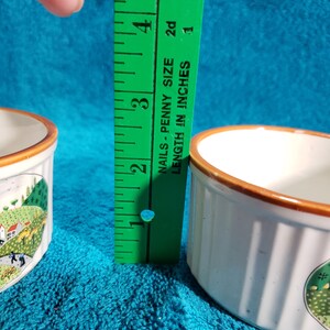 May include: Two white ceramic ramekins with brown rims and a country scene design. The ramekins are 1 1/2 inches tall.