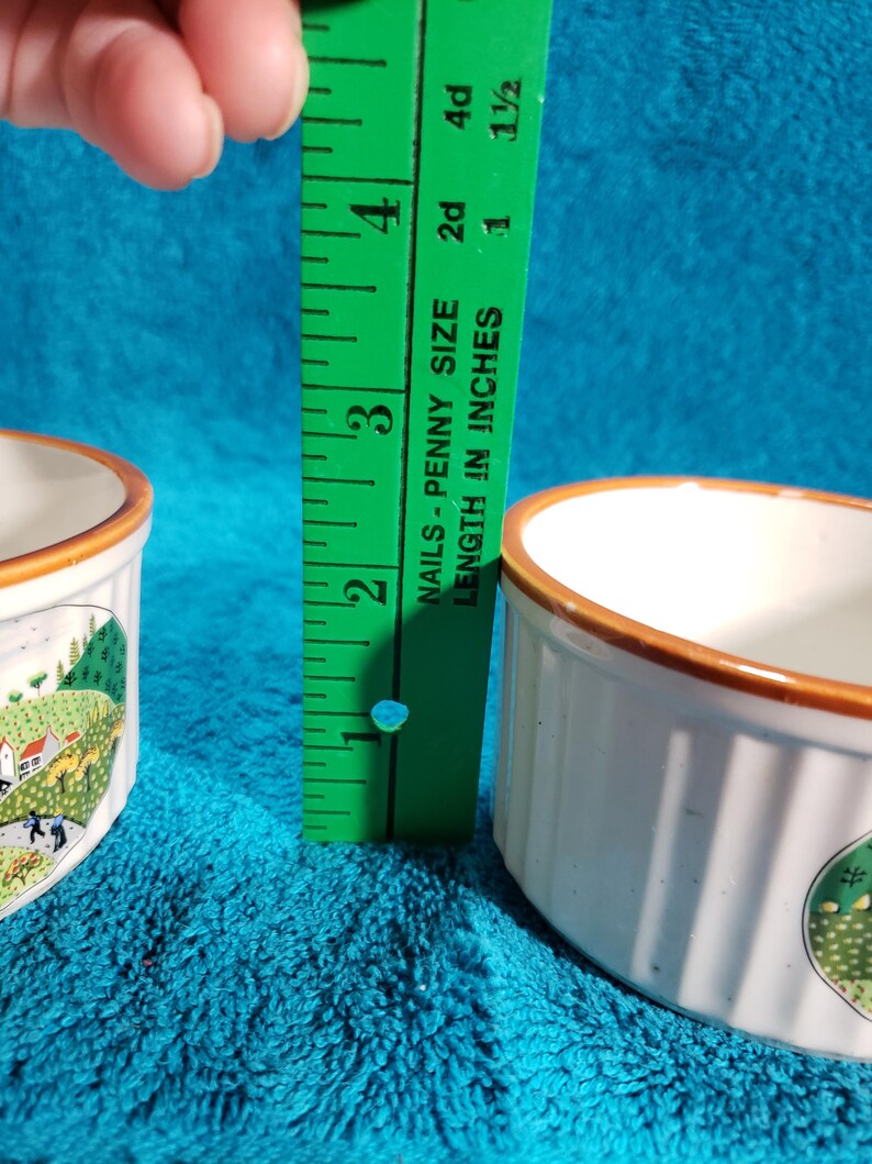 May include: Two white ceramic ramekins with brown rims and a country scene design. The ramekins are 1 1/2 inches tall.