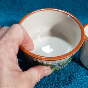 May include: Set of four white ceramic ramekins with a brown rim and a colorful countryside scene printed on the outside.