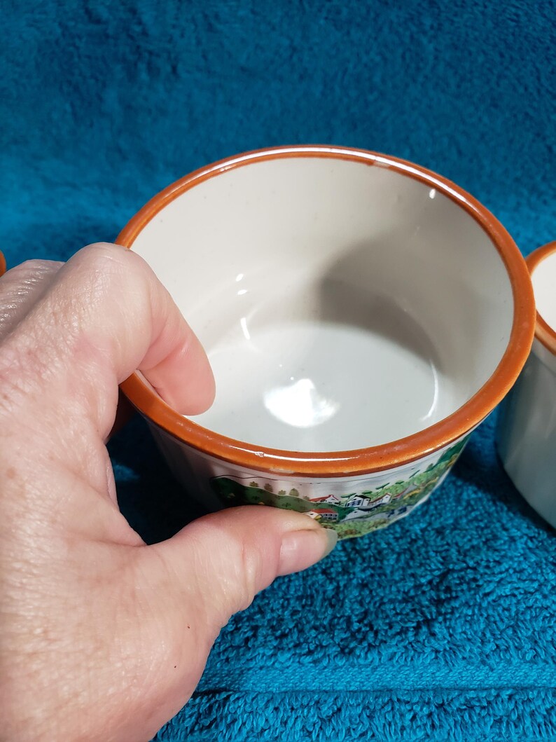 May include: Set of four white ceramic ramekins with a brown rim and a colorful countryside scene printed on the outside.
