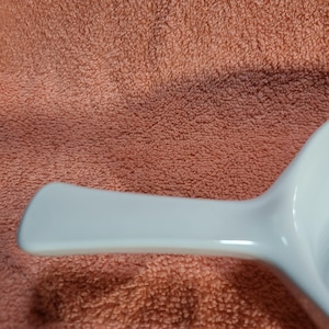 May include: A white, rectangular-shaped serving dish with a handle. The dish is made of a glossy material and is set against a peach-colored, textured background. The handle is smooth and curved.