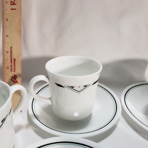 May include: Set of four white ceramic teacups and saucers with a black and green trim. Each cup features a small pink flower design.