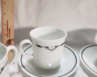 May include: Set of four white ceramic teacups and saucers with a black and green trim. Each cup features a small pink flower design.