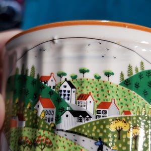 May include: A white ceramic ramekin with a colorful illustration of a small town with houses, trees, and a winding road. The illustration is surrounded by a light orange rim.