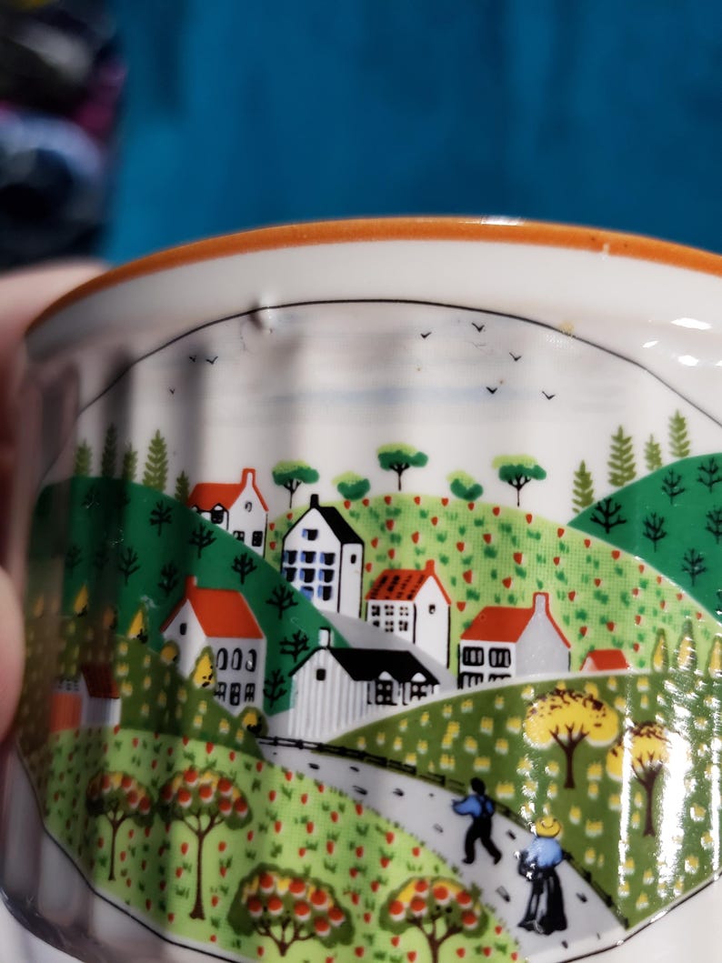 May include: A white ceramic ramekin with a colorful illustration of a small town with houses, trees, and a winding road. The illustration is surrounded by a light orange rim.