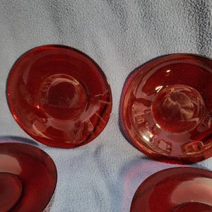 May include: A collection of ruby red glass saucers. The saucers have a circular shape with a slightly raised center and a smooth, glossy finish. The color is a deep, rich red, and the glass appears to be transparent.