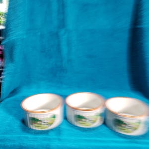 May include: Three white ceramic mugs with a brown rim and a green and white landscape design. The mugs are on a blue surface.