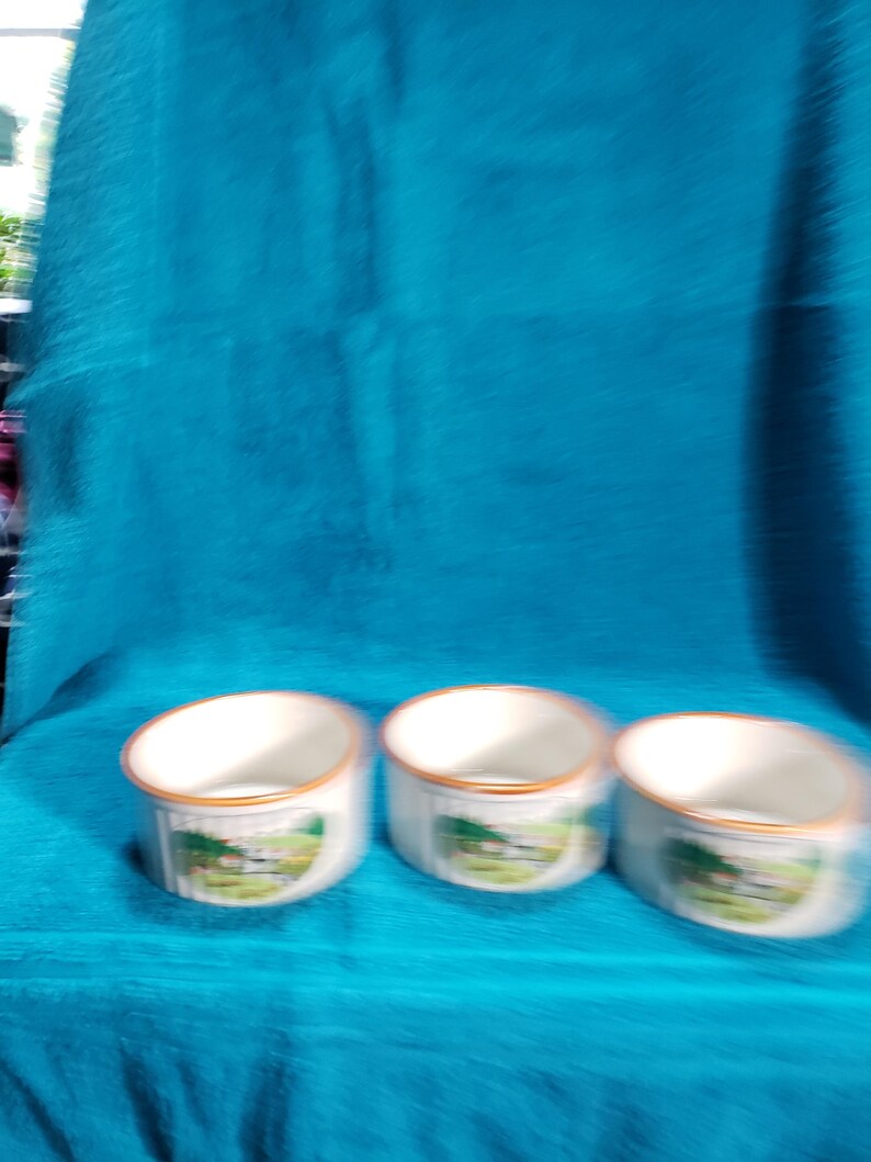 May include: Three white ceramic mugs with a brown rim and a green and white landscape design. The mugs are on a blue surface.