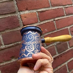 Könnte beinhalten: Eine marineblaue und kupferfarbene Kaffeekanne mit Holzgriff. Die Kanne hat ein verziertes Design in Kupfer. Der Griff ist hellbraun. Die Kanne wird vor einer roten Backsteinmauer gehalten.