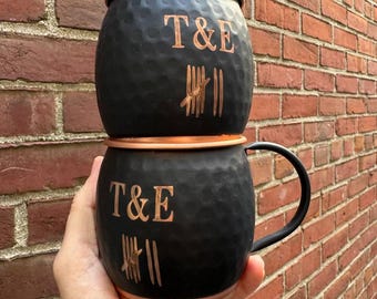 May include: Two black Moscow Mule mugs with copper rims and lids. Each mug has the letters "T&E" and vertical lines etched in copper. The mugs are stacked, and the background is a brick wall.