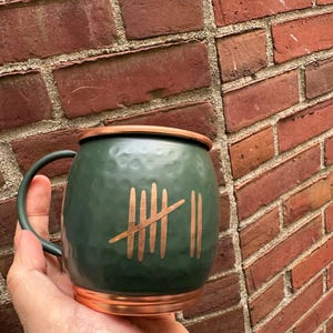 May include: A green hammered copper mug with a copper rim and a gold tally mark design. The mug is in the palm of a hand.