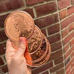 May include: A set of four circular copper coasters featuring a distinct hammered texture on the surface. Each coaster has a raised rim and a polished metallic finish that reflects light, showcasing the warm, reddish-brown tone of the copper material.