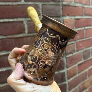 May include: A copper-coloured Turkish coffee pot with an etched floral design. The pot has a wooden handle and a flared rim. The etched design features intricate patterns. The pot is held against a brick wall background.