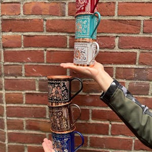 Op de afbeelding: Een stapel van zes metalen mokken met ingewikkelde bloemengravures en koperkleurige randen. De mokken zijn rood, groenblauw, wit, zwart, brons en blauw van kleur, elk met een bijpassend handvat en een gedetailleerd patroon van bloemen.