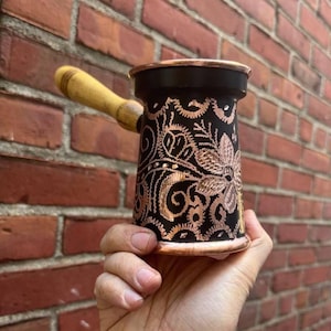 May include: A black and copper Turkish coffee pot with a wooden handle. The pot features intricate floral and swirl designs in copper against a black background. The pot is held in a hand.