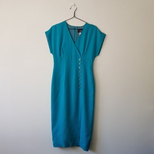 Puede incluir: Un vestido azul turquesa con mangas cortas y escote en V. El vestido tiene una fila de botones dorados en el lateral.