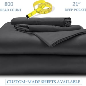 Puede incluir: Un juego de sábanas de cama de color gris oscuro con un conteo de hilos de 800 y bolsillos profundos de 53 cm. Las sábanas están dobladas y apiladas sobre un fondo blanco. Sábanas hechas a medida disponibles.