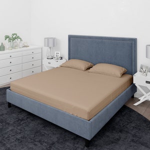 May include: A tan fitted sheet on a blue upholstered bed frame with a headboard. The bed is made with two tan pillowcases.