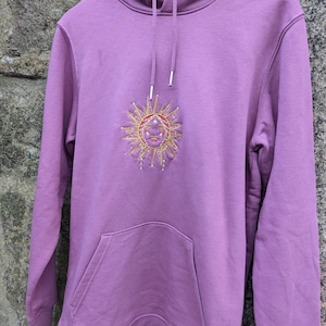 May include: Mauve hoodie with a front pocket and drawstring hood. The hoodie features an embroidered sun design in gold and red thread. The sun has a smiling face and radiating rays.