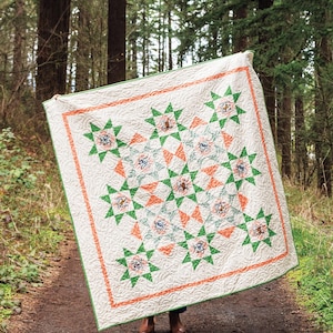 May include: A cream-colored quilt with a green and orange star pattern. The quilt has a green border and a detailed quilted design. The quilt is held up in a forest setting, showcasing its handmade quality and design.