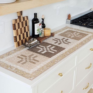 Può includere: Un runner da tavola trapuntato rettangolare con un motivo geometrico ripetuto nei toni del beige, marrone chiaro e marrone scuro. Il runner presenta un bordo decorativo con motivo floreale ed è appoggiato su un piano cucina bianco.