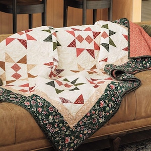 Könnte beinhalten: Eine Patchwork-Decke mit cremefarbenem Hintergrund und sich wiederholenden Sternmustern in Rot-, Grün- und Brauntönen. Die Decke ist mit einem dunkelgrünen Rand mit Blumenmuster versehen und weist eine texturierte, gesteppte Oberfläche auf.
