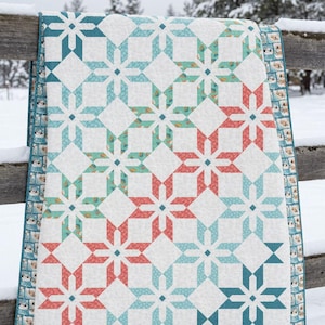 May include: A quilt featuring a repeating star pattern in shades of coral, teal, and light blue against a white background. The quilt has a border with a small-scale print of snowmen and is stitched with a textured, quilted finish throughout.