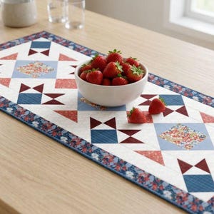 May include: A rectangular table runner with a geometric pattern in red, blue, and white. The runner features a floral border and a bowl of fresh strawberries. The runner is placed on a light-colored wooden table.