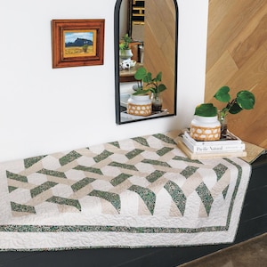 May include: A geometric quilt with a repeating pattern of green and beige shapes on a white background. The quilt is placed on a dark surface, with a small painting and a black arched mirror in the background. A decorative jar and books are also visible.