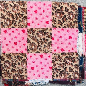 May include: A square quilt block featuring a nine-patch grid pattern. The design alternates between squares of tan fabric with a dark brown floral print and squares of pink fabric with a small red leaf pattern, creating a checkerboard effect.