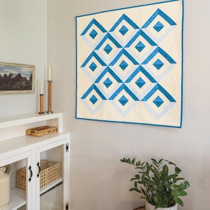 May include: A blue and white geometric quilt with a diamond pattern hangs on a white wall. The quilt is framed with a white border. A white cabinet with glass doors and shelves is in the corner of the room. A potted plant sits on a wooden table.