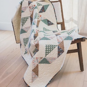 May include: A quilted blanket featuring a geometric pattern of cream-coloured fabric interspersed with triangular patches in shades of green, brown, and floral prints. The quilt has intricate swirling stitch details and a dark, patterned fabric border.