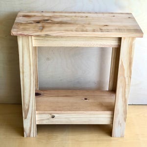 May include: A simple, light wood end table with a single shelf. The table has a natural wood finish and is made of solid wood.