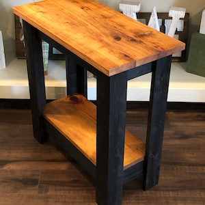May include: A small wooden side table with a natural wood top and black painted legs and shelf. The table has a simple design with a rectangular top and a single shelf.